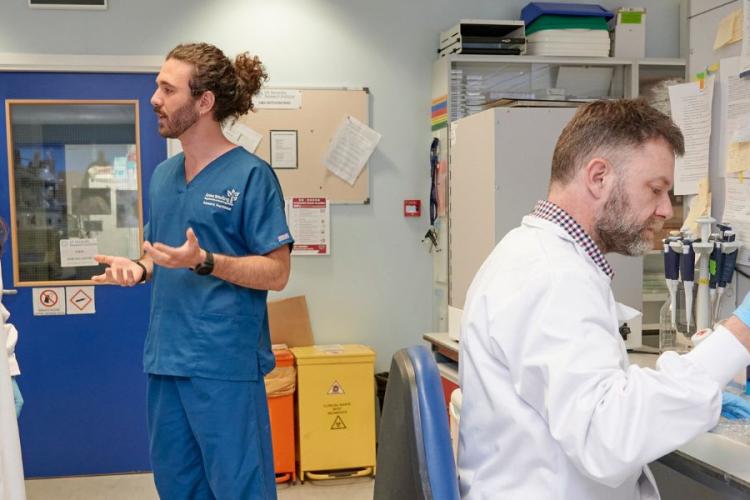 Inside of a busy laboratory, in the background an Anne Clinic researcher and a laboratory scientist and are in a discussion. In the foreground, a scientist is using a pipette at the lab bench.  