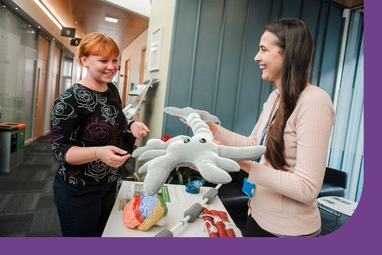 A picture taken inside the Anne Rowling Clinic reception area. A researcher is holding a plush-toy neuron to show to a member of the public who is attending a Research Open Evening