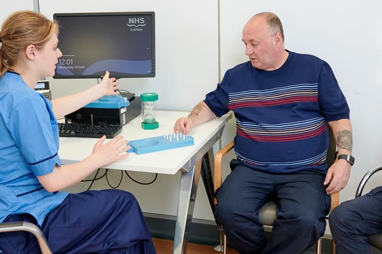An NHS research practitioner is in a clinic room with two men. One is performing a cognitive test, moving pegs on a plastic board, while the other looks on.