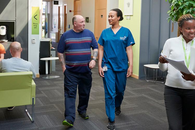 A picture taken inside the Anne Rowling Clinic reception area, people are sitting waiting for their appointment, a researcher is talking and walking with a patient and two staff members are chatting