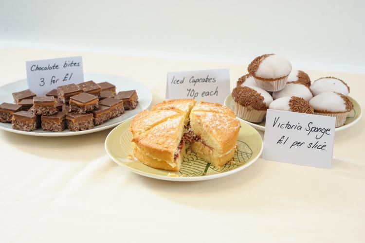 Image of a bake sale with chocolate bites (right), Victoria sponge cake (centre) and chocolate cupcakes (left)