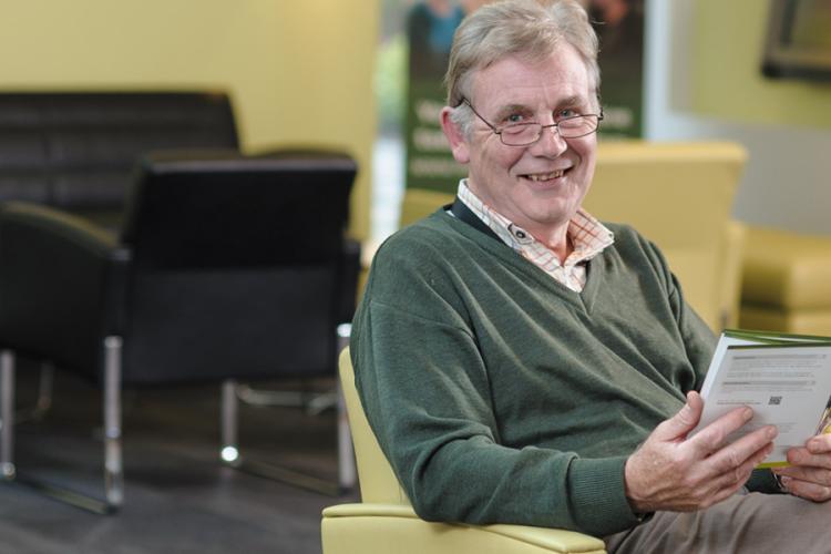 An older person is pictured sitting in Anne Rowling Clinic reception area, smiling at the camera whilst holding a leaflet