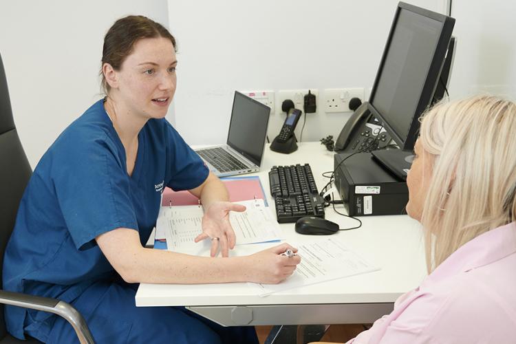 A research nurse and a patient are in discussion in a clinic room, the nurse is making notes