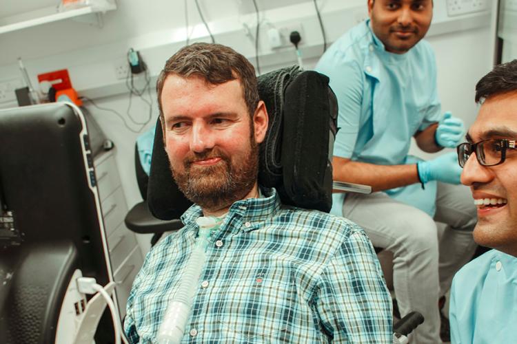 Two researchers in pale blue lab coats are talking happily in a lab setting with a man who is sitting in a power wheelchair and looking at a screen 
