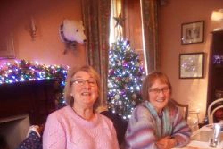 A picture of fundraiser Linda McTurk at a table laden with lots of cakes and baking, the setting is Christmas and there are sparkling lights in the background. Linda and her friend are smiling at the camera while enjoying a cup of tea.