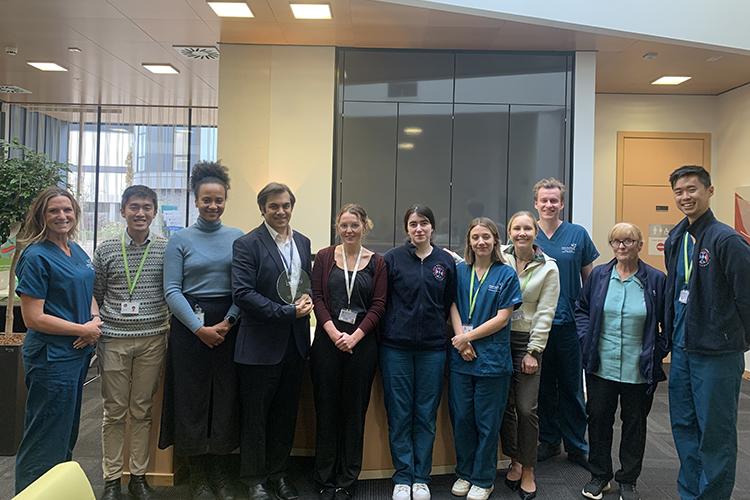 A group of 11 people, standing smiling in a line in the reception area of the Anne Rowling Clinic. The team lead is holding a glass award.