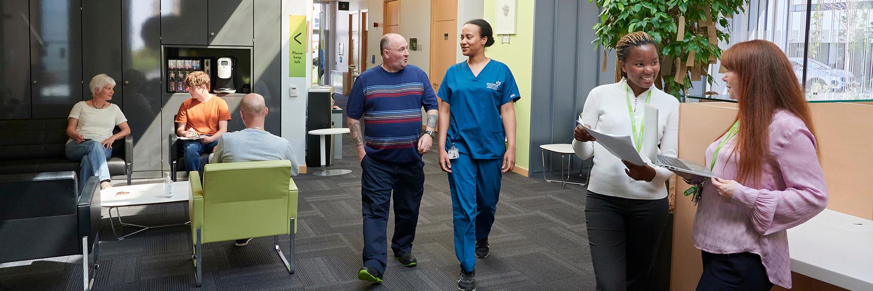 A picture taken inside the Anne Rowling Clinic reception area, people are sitting waiting for their appointment, a researcher is talking and walking with a patient and two staff members are chatting