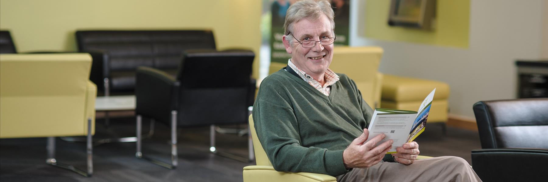 An older person is pictured sitting in Anne Rowling Clinic reception area, smiling at the camera whilst holding a leaflet