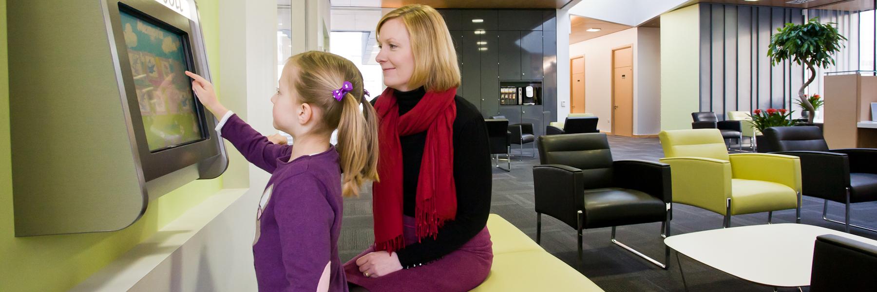 A woman watching and smiling while her young daughter plays on the "Magic Wall"; wall-mounted touchscreen device.