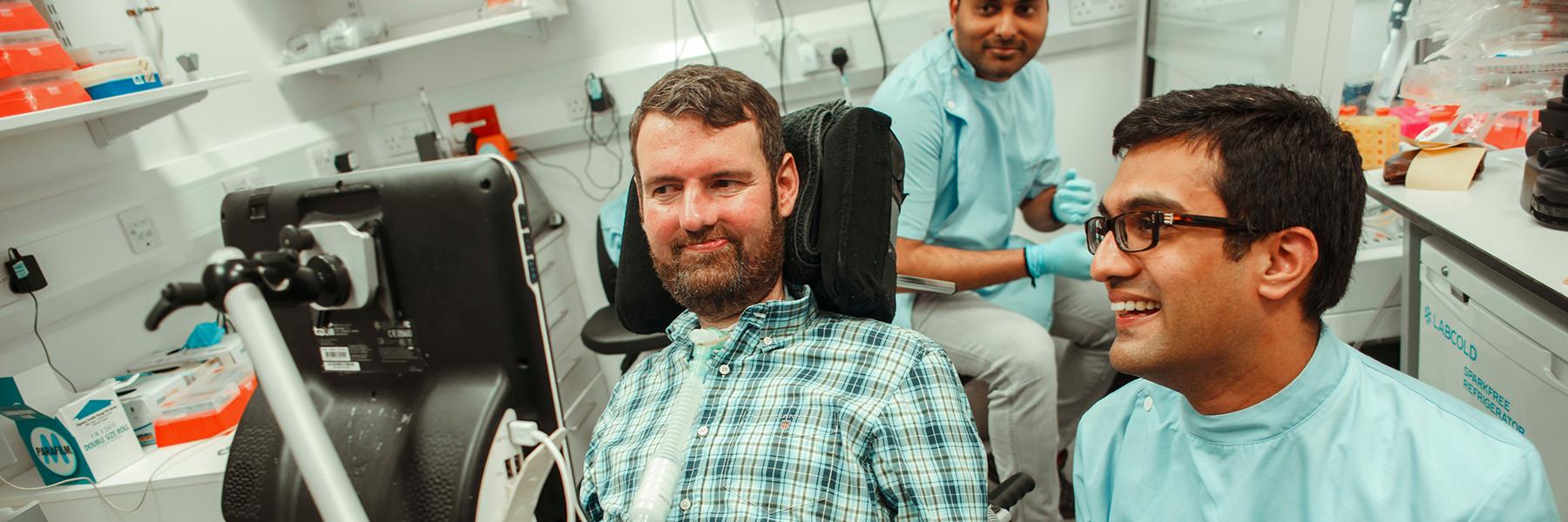 Two researchers in pale blue lab coats are talking happily in a lab setting with a man who is sitting in a power wheelchair and looking at a screen 