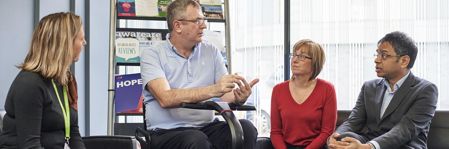 A patient in a wheelchair and their carer are in discussion with researchers