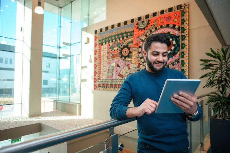 A person smiling and reading from an electronic device with colourful tapestry in the background